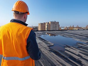 Roof-Maintenance-San-Diego-CA-California-2 | Sully-Jones Roofing Construction worker wearing an orange safety vest and hard hat inspecting a flat rooftop with puddles, city buildings in the distance.